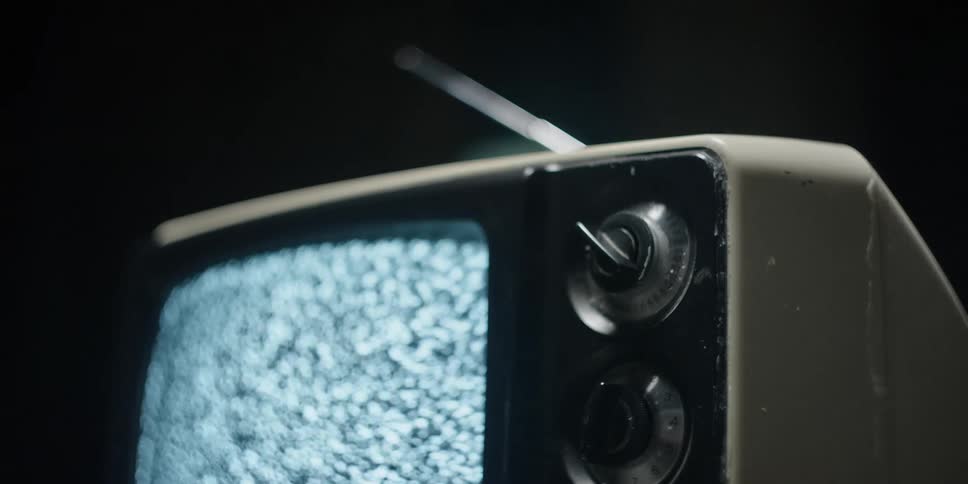 The screen of an older cathode ray television set is showing only static as someone reaches out to turn one of its knob