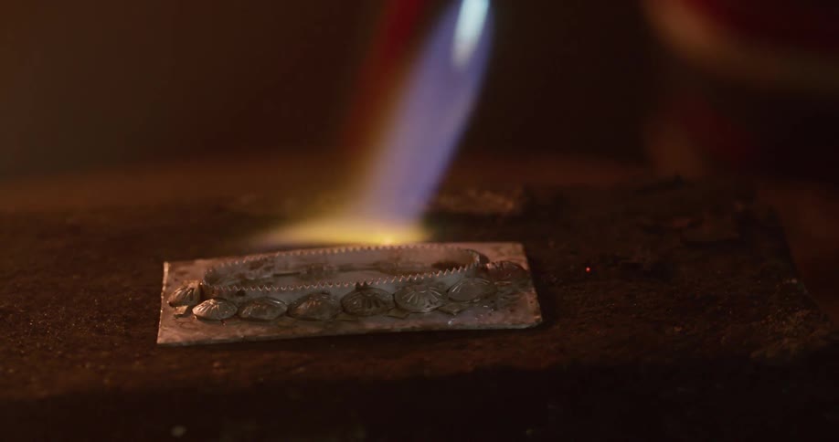 Blue flame flows from torch heating silver design on rotating fireproof block