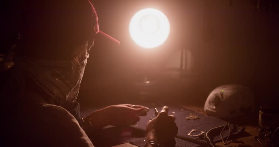 A person with a hat and a handkerchief over their face sitting at a table using tools