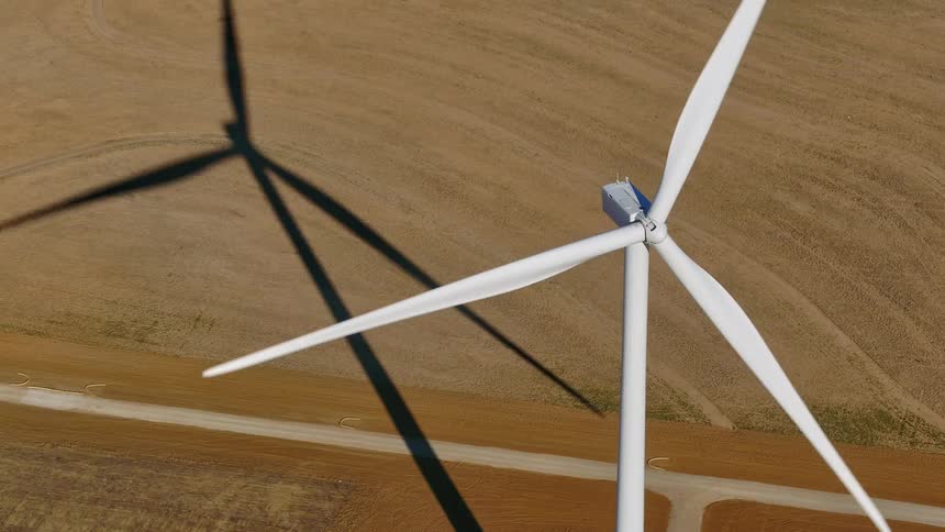 White wind turbine stands on yellow green field casting shadow across road and nearby fields