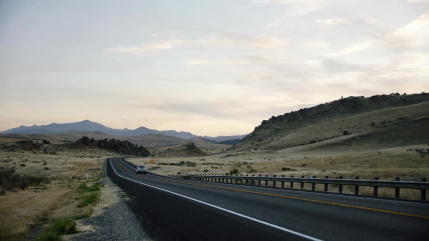 A grey pickup truck drives on highway through a field at dusk