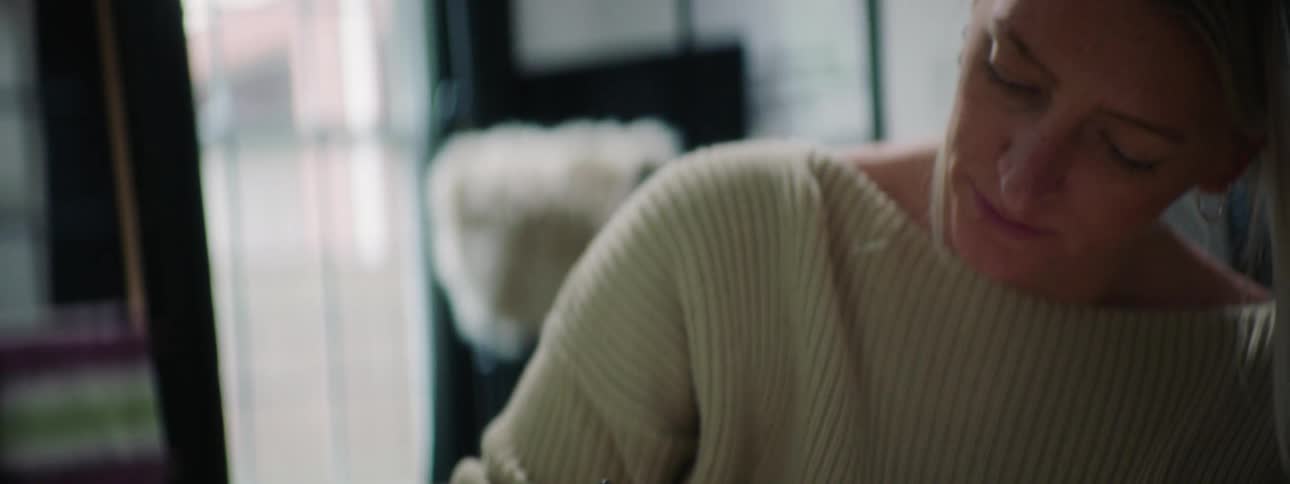 Blonde woman with earrings and white sweater sitting on the sofa writes in a notebook in the living room