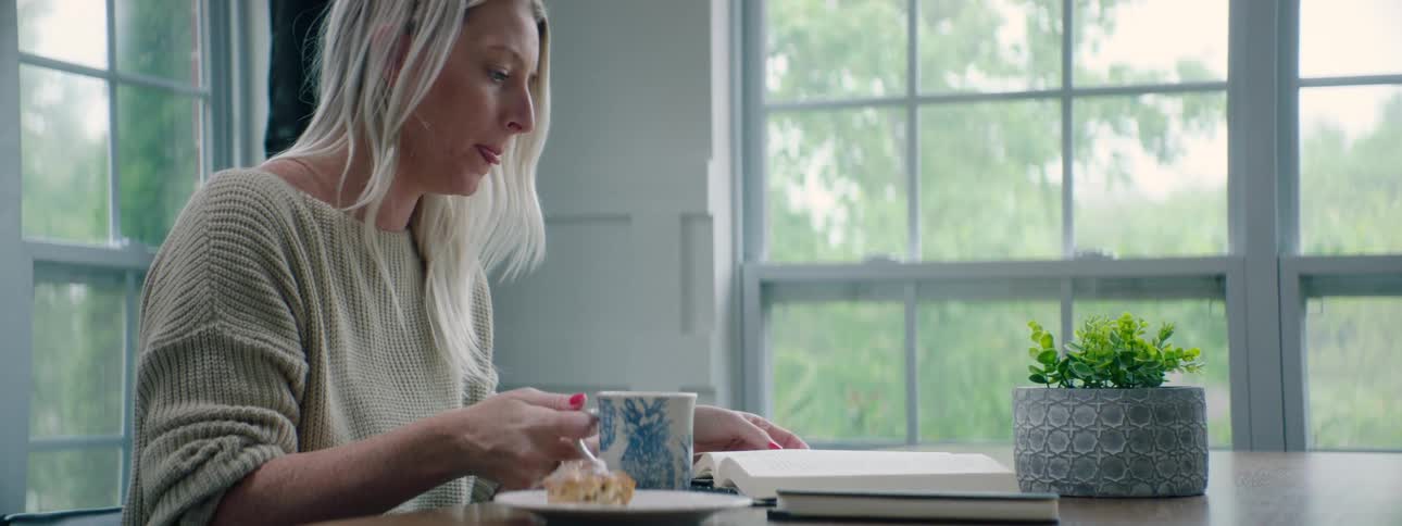 A woman sits at a table reading a book eating her breakfast and drinking a beverage from a mug