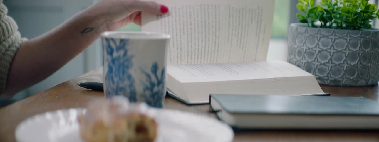 A woman sitting at a table sits down a coffee mug before turning the page of a book lying the table