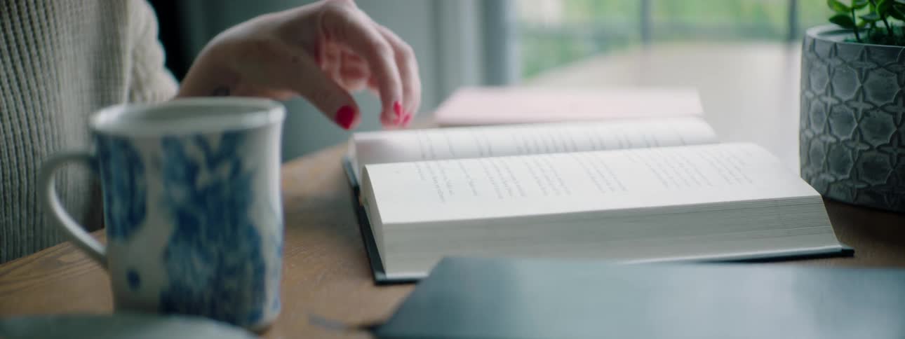 A woman sitting at home reads a book as a snack and a cup of coffee sit beside her