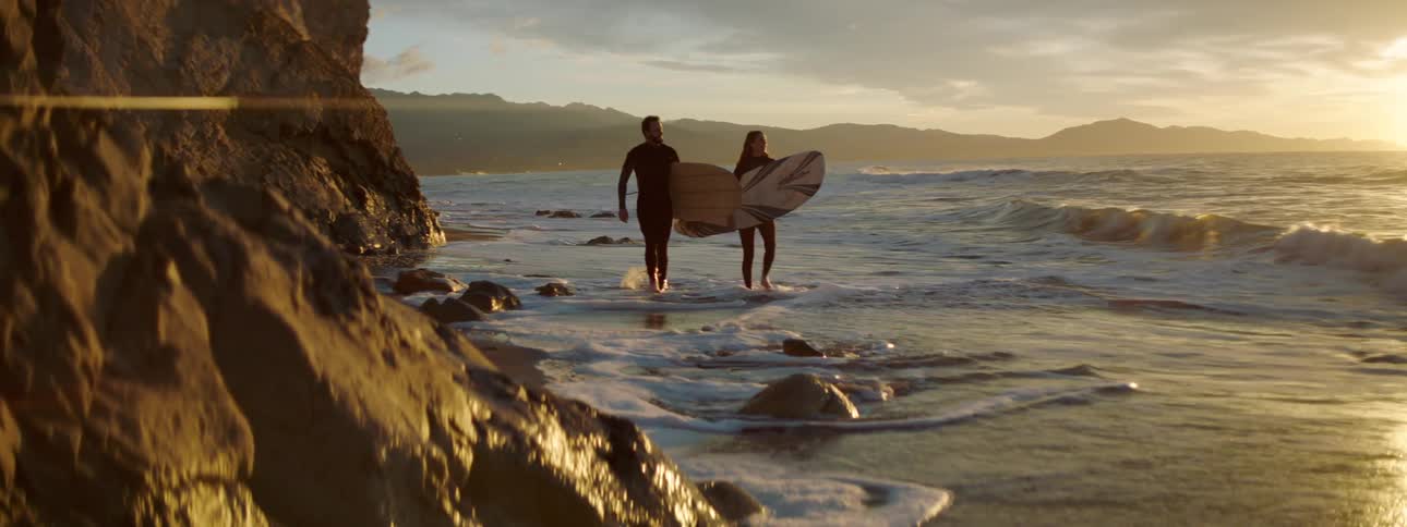 A pair of surfers carrying surfboards walk along a beach beneath a cliff in the evening