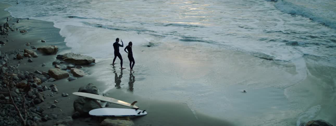 A man and woman stand on a beach wearing wet suits with surfboards lying behind them on the sand