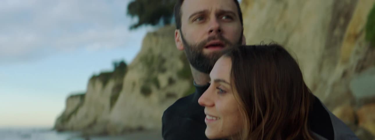 A man and woman in wetsuits embrace while chatting and laughing on the beach