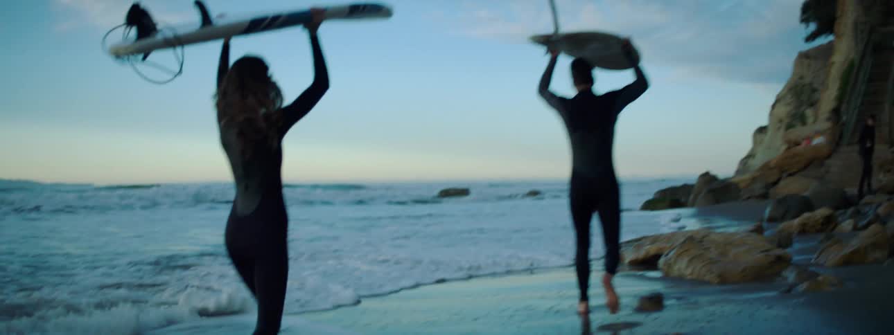 A man and woman run along the shore carrying surfboards
