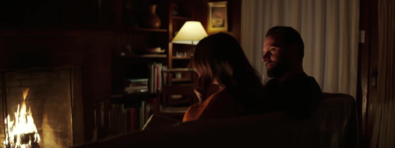 Man with a beard puts his arm around womans shoulder and watch a fireplace together in the living room