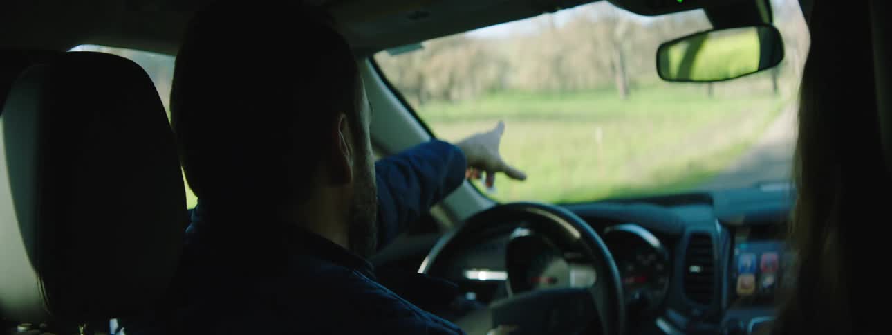 Man with a beard talks to a woman with brown hair while driving on a country road