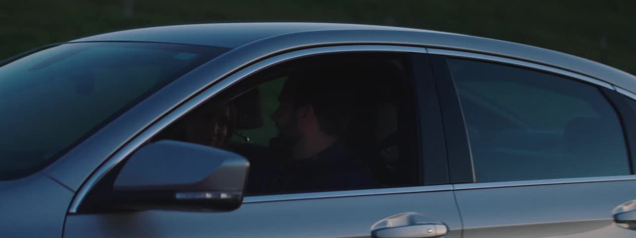 A man driving a car through the countryside points at something out of his window for his female passenger
