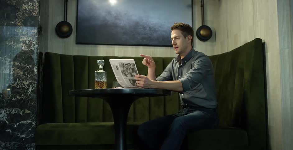 Man sitting at a table in a bar reads a newspaper next to a liquor bottle