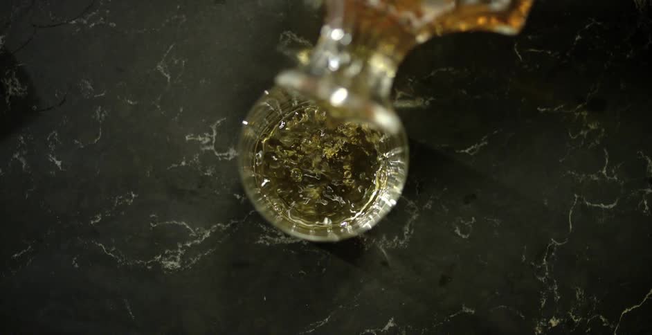 An amber colored liquid is poured into a drinking glass on a marble counter