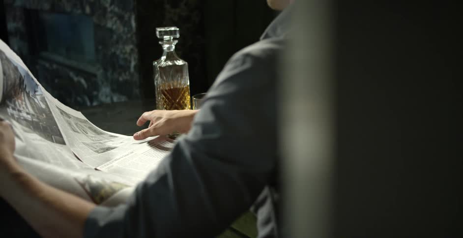 A man sits at a table with a bottle of liquor and drinking glass on it while reading a newspaper
