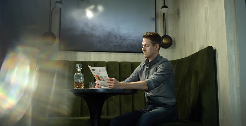 A man sitting in a booth at a bar reads a paper with a drink on the table