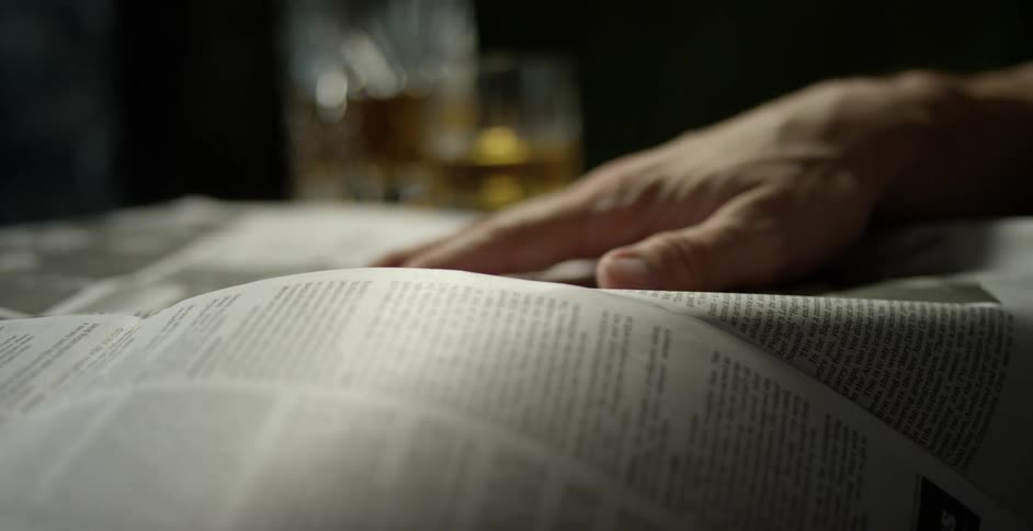 A man holds down a newspaper with his hand as he reads it at a table