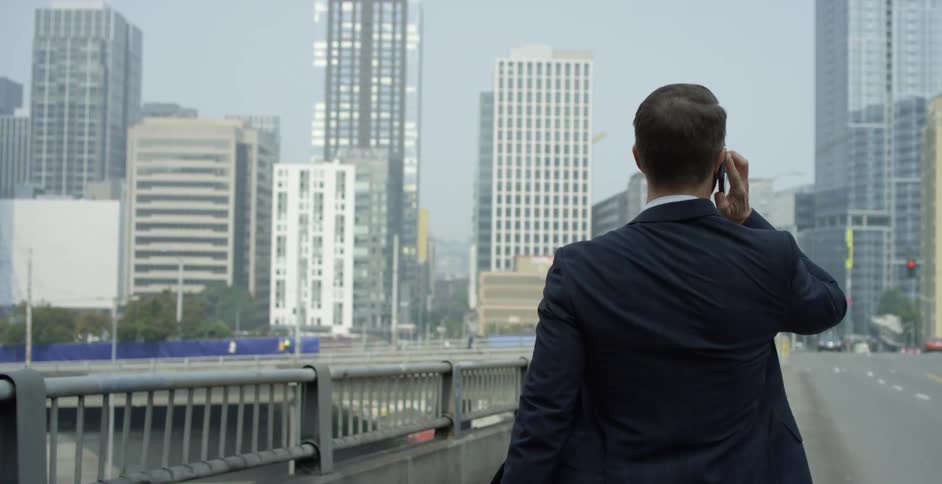 Man with a black suit and sunglasses crosses a bridge while talking on a smartphone