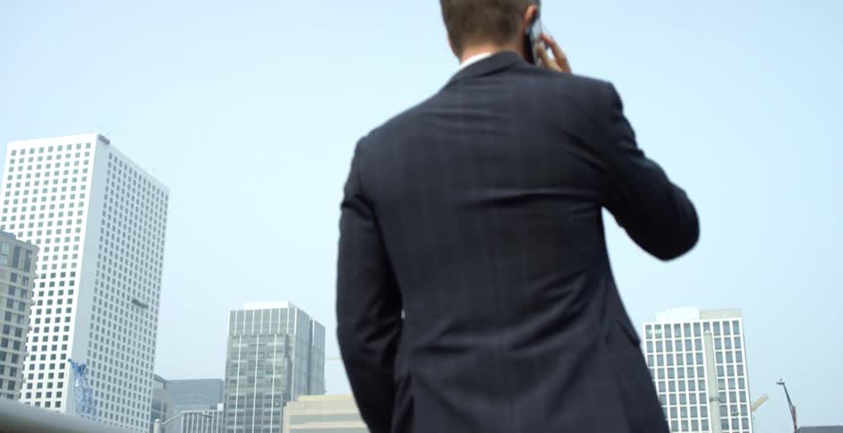 Businessman with a black suit talks on a smartphone while crossing a bridge in a city