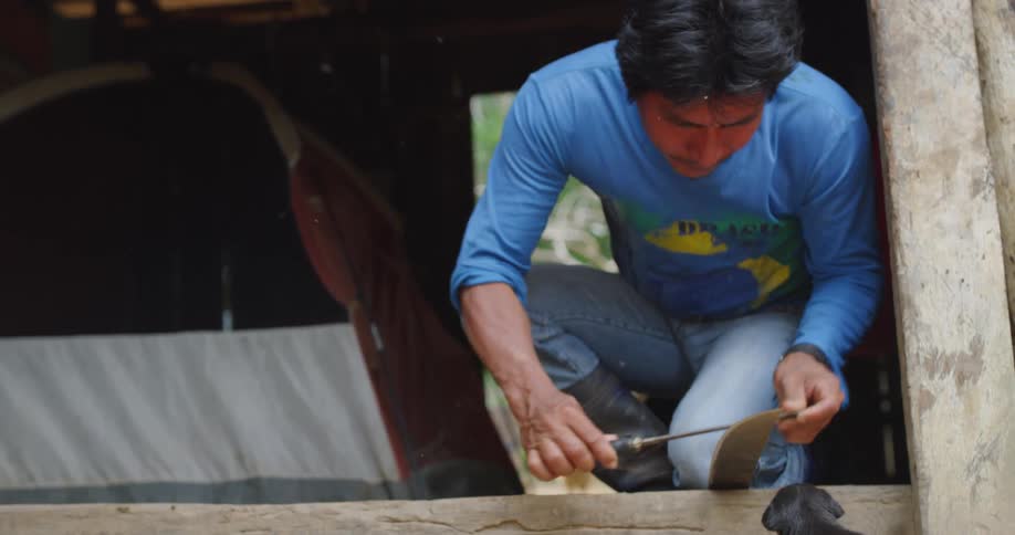 A man sharpens a machete next to a curious black puppy