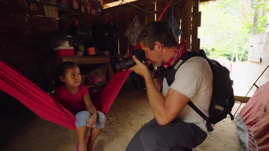 A photographer kneels to take a photograph of a little girl sitting on a hammock in a wooden shack