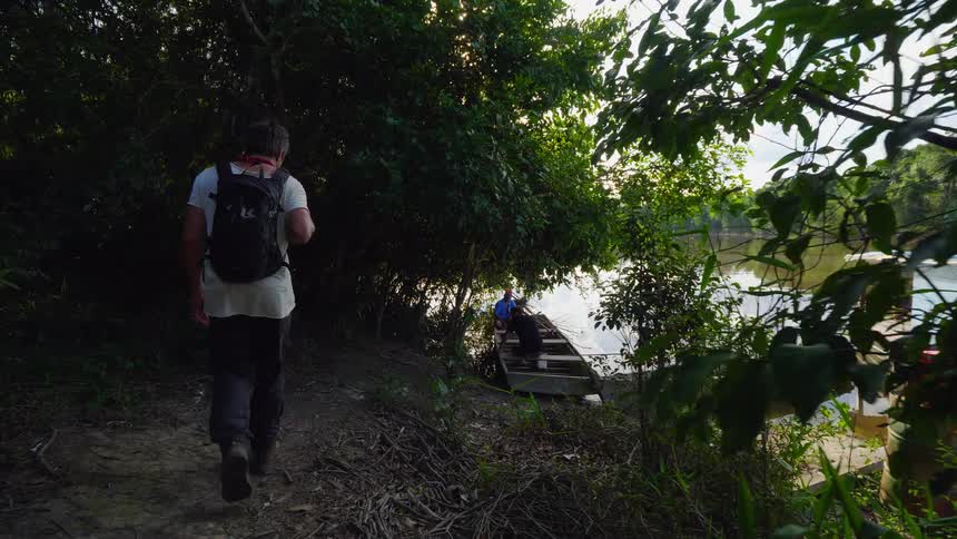 A man walks along a dirt path through a forest towards a small boat docked on the river