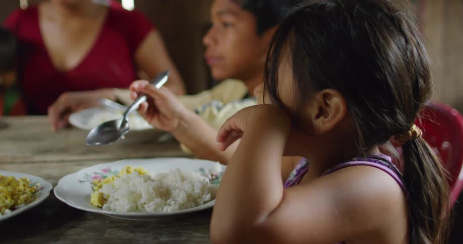 A little girl holds a spoon as she sits at a wooden table next to her brother eating