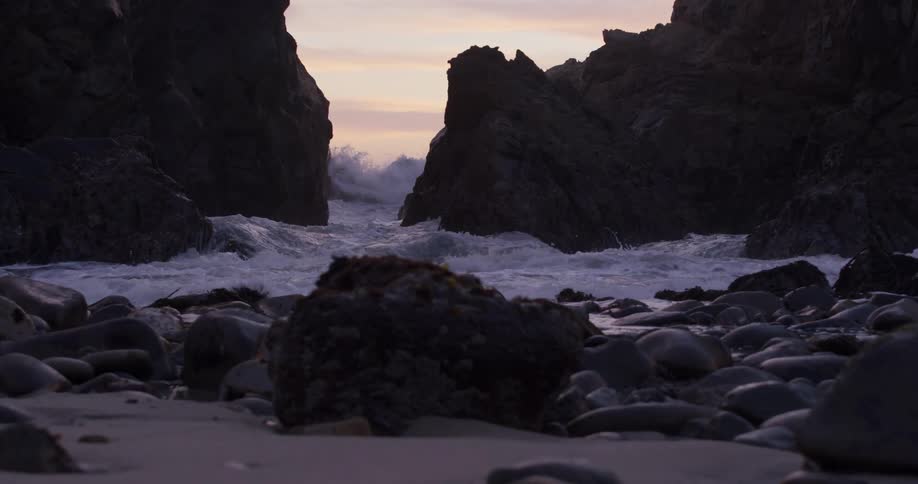 Foamy ocean waves crash against big rocks at dusk