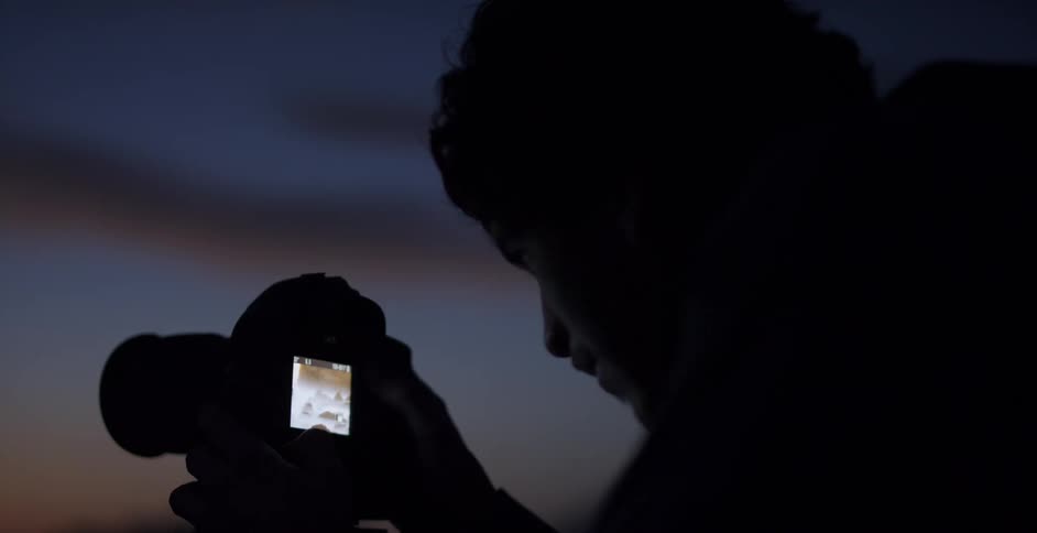 A photographer looks at the screen of his camera on a tripod and talks and gestures with his hands