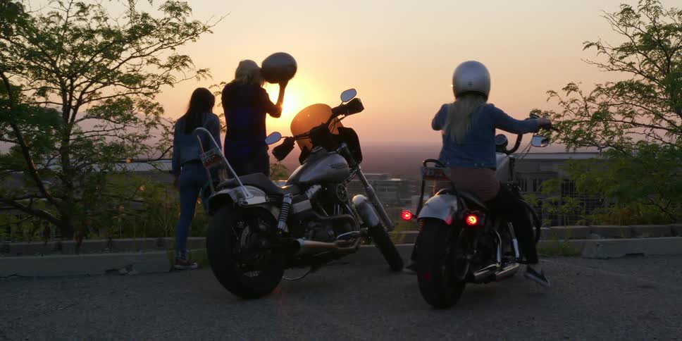 Three woman on two motorcycles with helmets with the sky and the sun in the background