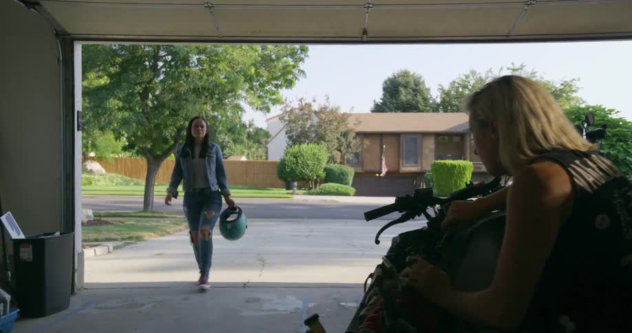 Person works on motorcycle in garage as girl walks in carrying helmet