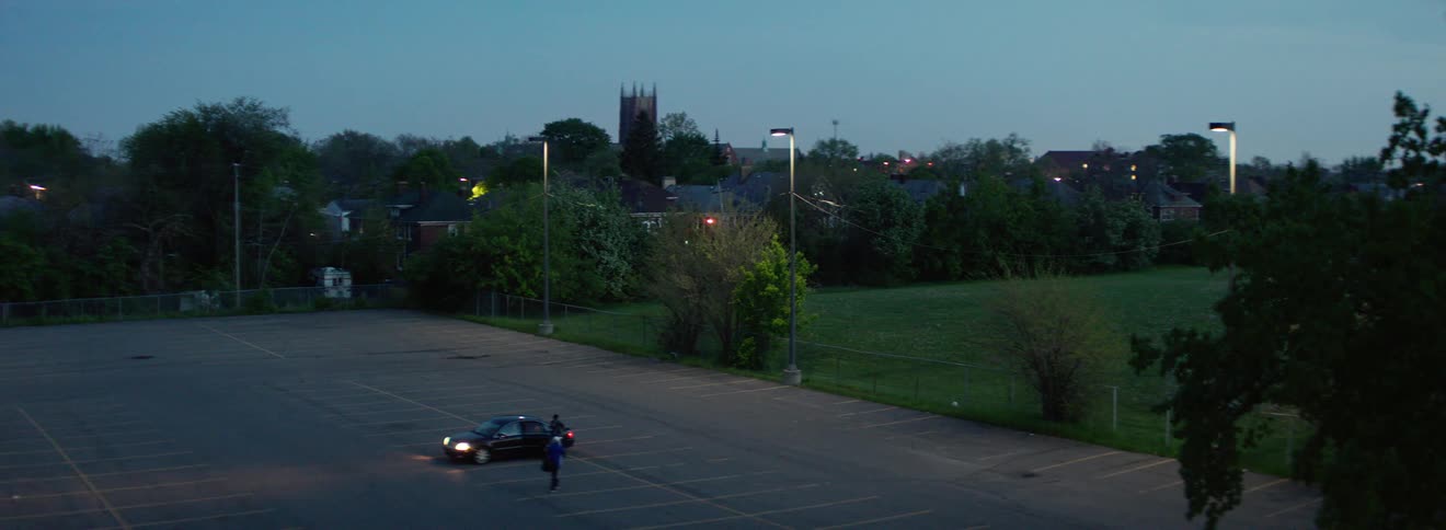 A car parked in a parking lot where a person enters into it and another person opens the trunk with buildings and the sky in the background
