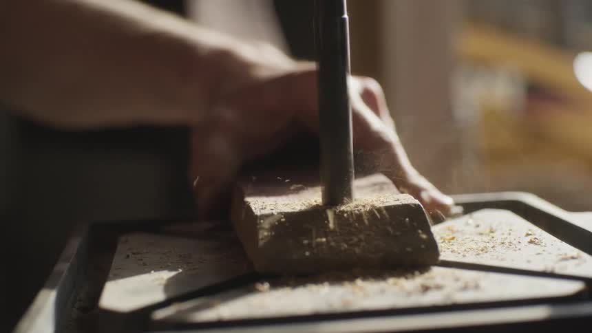 A man holds a piece of wood in place with his hand on a drill press as he drills into it