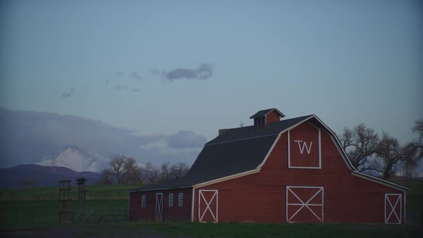 A barn with a vehicle passing in the foreground and trees mountains and the sky in the background