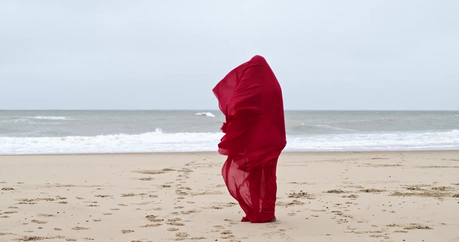 Blonde woman wearing a red scarf stands on the beach and watches the sea on a cloudy day