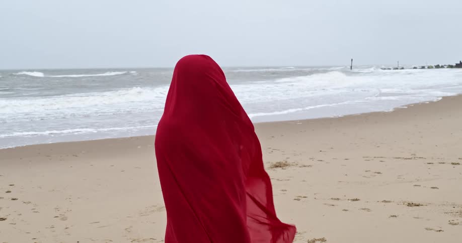 Woman wrapped in a full body red veil stands on a beach in an overcast day