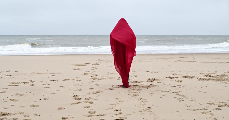 Blonde woman wearing a red scarf walks on the beach next to the sea on a cloudy day