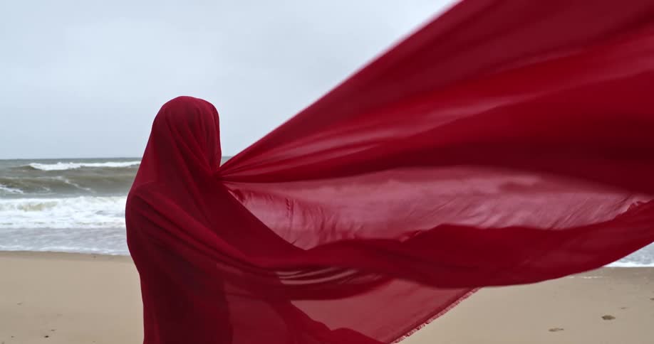 Person stands on beach wrapped in flowing red fabric as waves crash onto sand