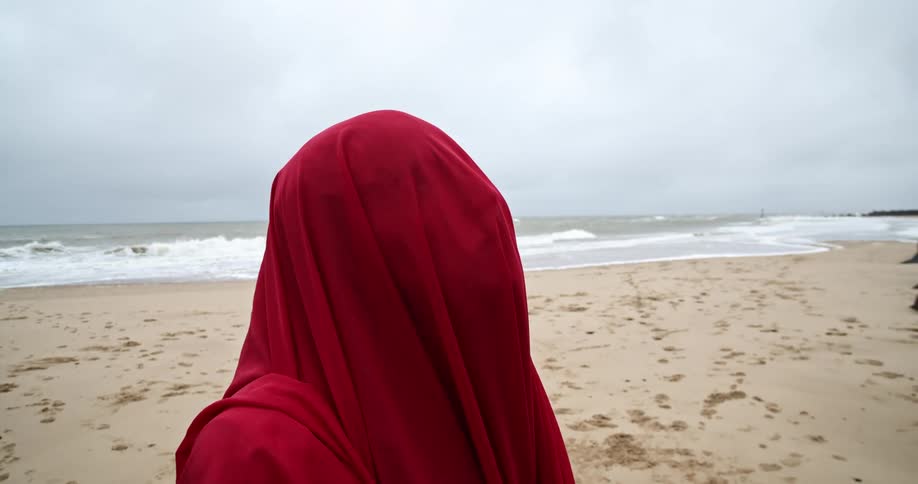 A person covered in a red robe stands on the beach