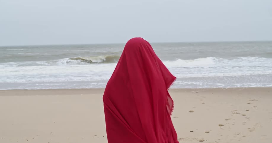 A person stands on a beach while hidden beneath a sheer red cloth that billows in the wind