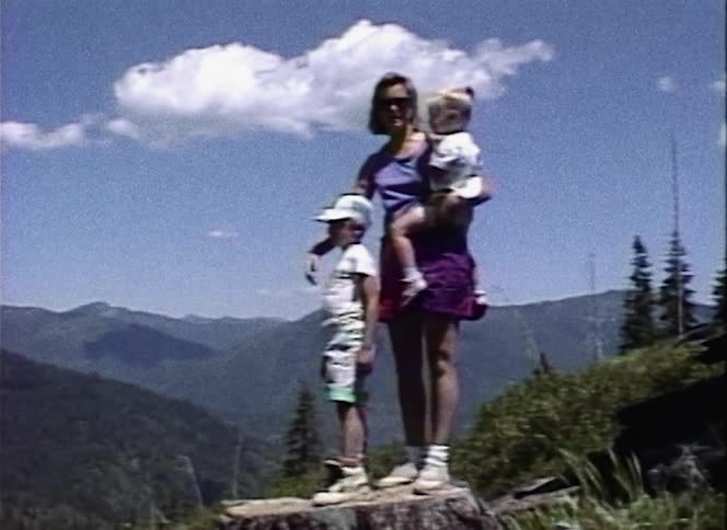 A mother and two children stand on a tree stump in a mountain forest