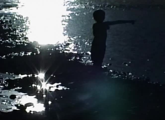 A silhouetted boy points while standing next to a lake