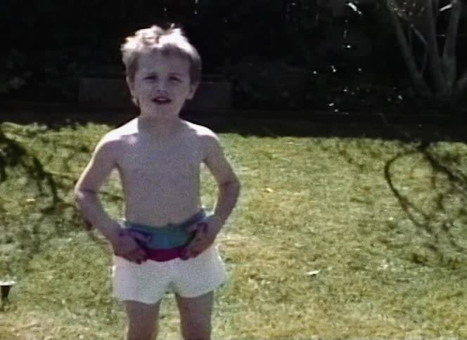 A boy speaks to the camera before running towards and standing on a surfboard in the back lawn of a house
