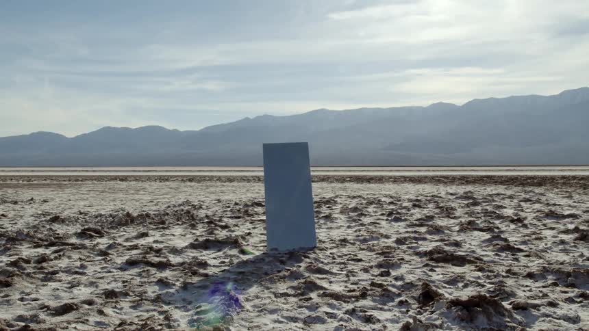 A steel slab in the middle of the desert with sand mountains the sky and clouds in the background