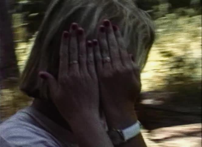 A woman walking in the woods covering her face with her hands with a tent and trees in the background