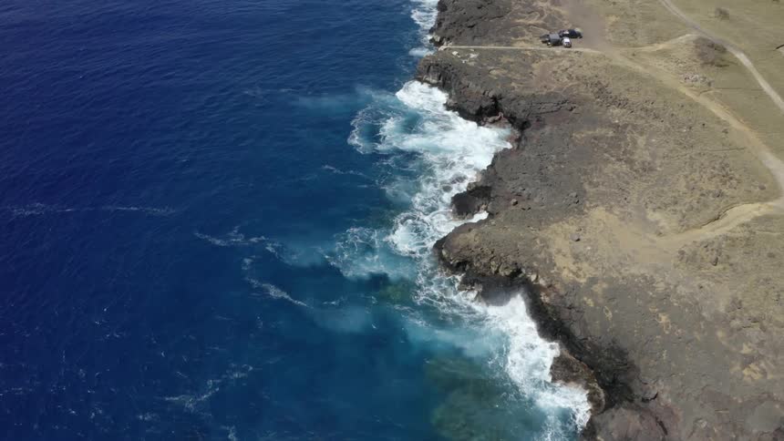 An aerial view of ocean waves crashing into a shoreline with parked cars