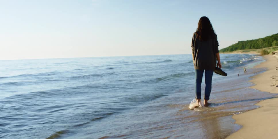 A woman walks along the sandy beach holding her flip flops