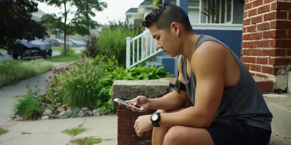 A teenager sits on the front step of a house and scroll on his cell phone
