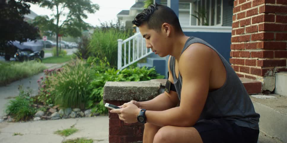 A man sitting down on steps in front of a house looking at a smart phone with trees and the sky in the background