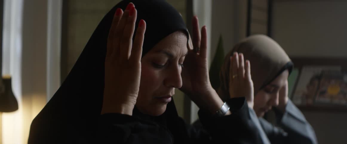 Two women with headscarves and loose garments perform the ritual prayers of islam in a room